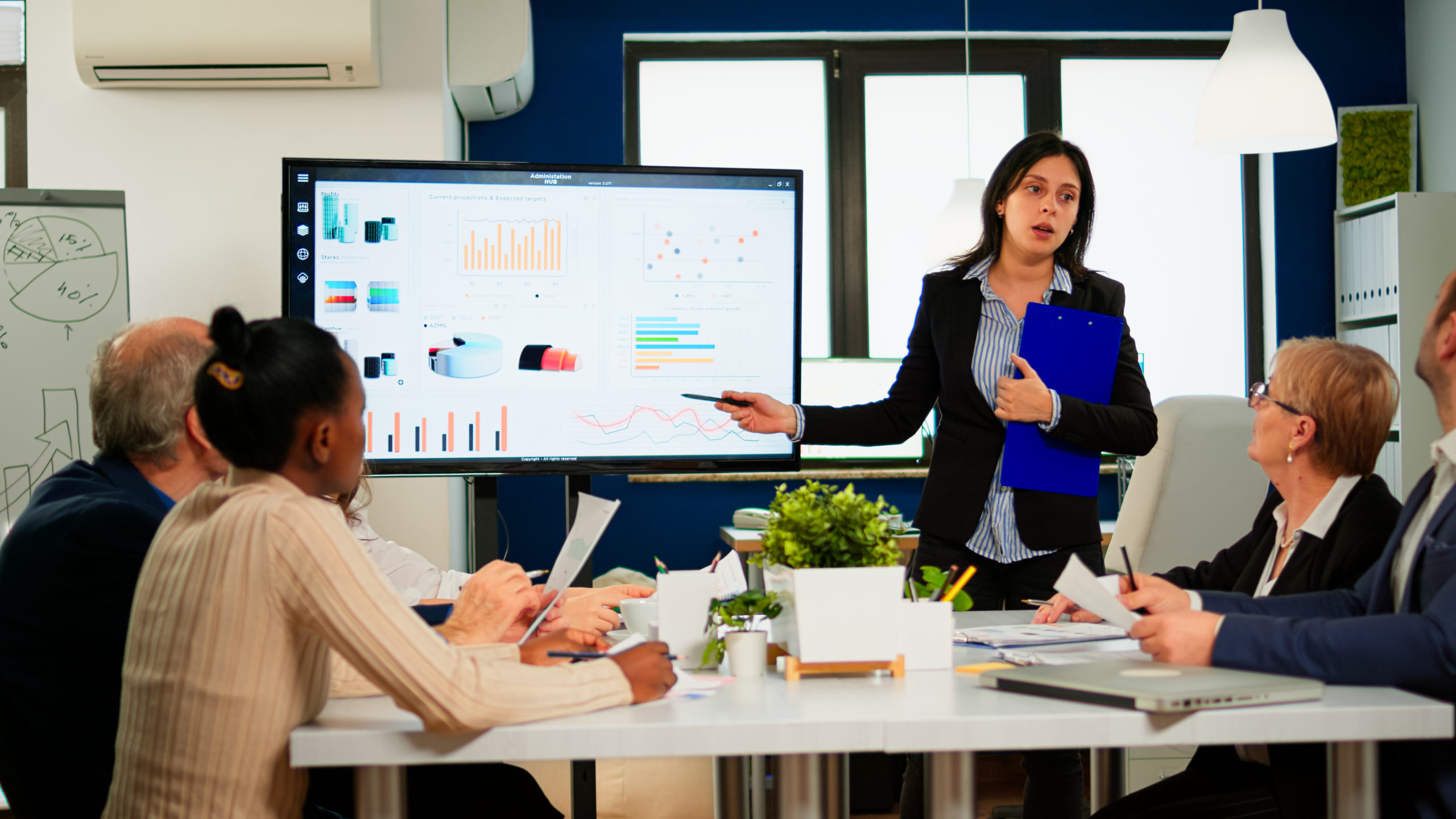 Female project manager holding financial meeting showing statistical graphs and charts on interactive whiteboard touchscreen device. Executive director working in broadroom of creative agency.